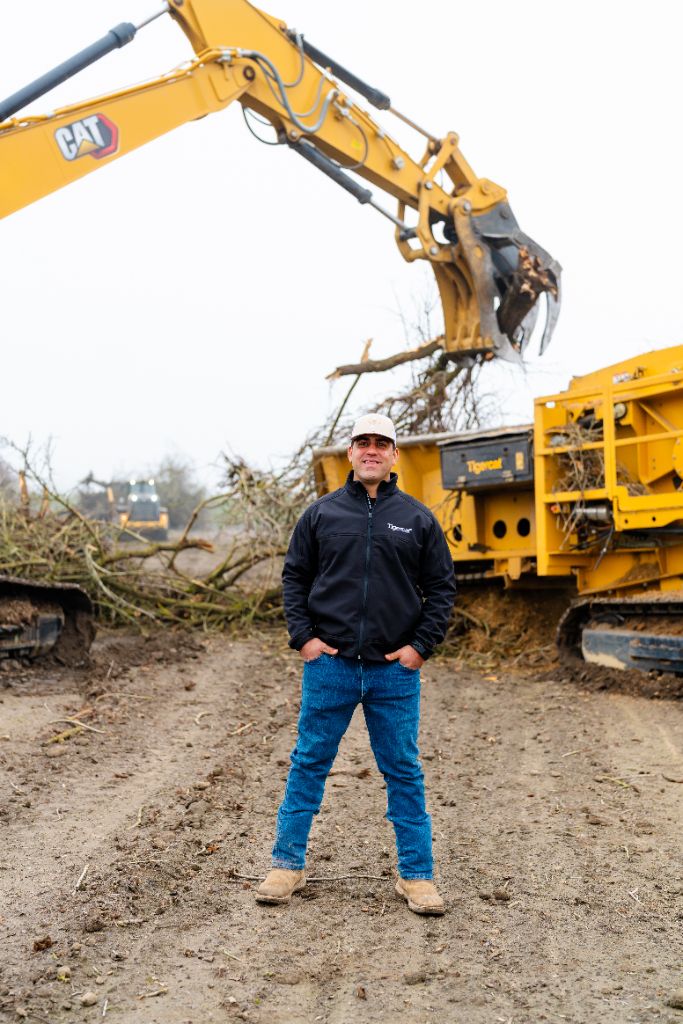 Anthony Nonini, owner of A. Nonini Ag Services, standing on an active orchard removal job with a CAT excavator and Tigercat grinder behind him